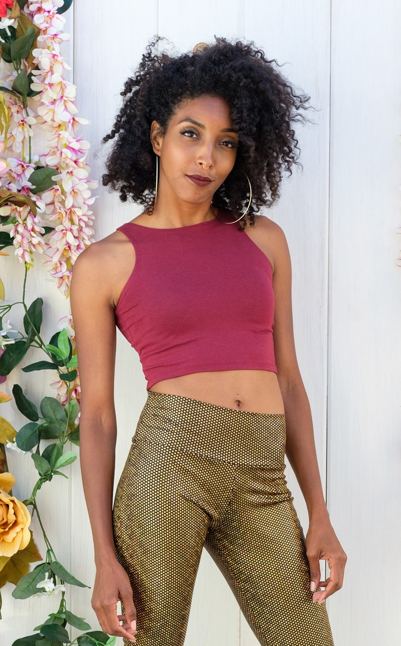May include: A woman with curly dark hair is wearing a burgundy crop top and gold patterned pants. She is standing in front of a white wall with pink flowers.