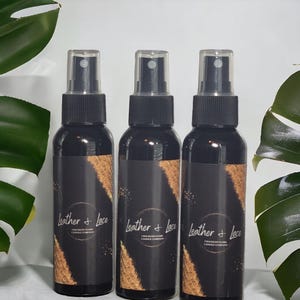May include: Three black spray bottles with clear tops are displayed. Each bottle has a black label with the words "Leather + Lace" and "Cracked Flame Candle Company" in white. The labels feature a gold and brown design.