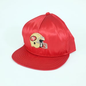 Puede incluir: Gorra de béisbol de satén rojo con un logotipo bordado de los San Francisco 49ers en oro y negro.