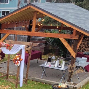 May include: Wooden outdoor structure with a grey tiled roof, string lights, and a decorative archway. Tables with burgundy tablecloths and beverage dispensers are set up for an event. Autumnal floral arrangements and pumpkins add seasonal decor.
