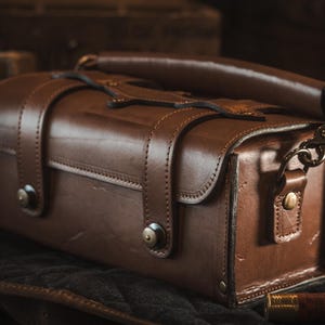 May include: A brown leather bag with a handle and shoulder strap. The bag has a rectangular shape with rounded edges and is secured with straps and metal hardware. The bag is likely for carrying items and has a vintage or rustic aesthetic.