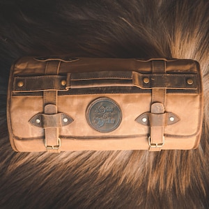 May include: A rectangular, tan canvas bag with brown leather accents, straps, and buckles. The bag features a handle and a circular emblem with the text "Sage & Quaker." The bag is set against a brown, textured background.
