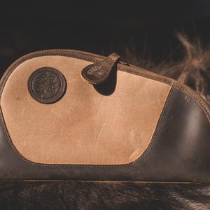 May include: A tan and brown canvas and leather case with a rounded shape. The case features a circular emblem and a leather pull tab. The case is resting on a textured, dark surface.