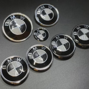 Puede incluir: Una colección de insignias con el emblema de BMW en varios tamaños. Las insignias presentan el logotipo de BMW, un diseño circular con segmentos negros y plateados, y las letras BMW. Las insignias están sobre un fondo oscuro.