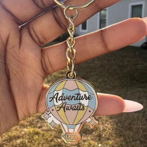 May include: A gold-colored keychain with a hot air balloon charm. The charm is round with a pastel color scheme and the words "Adventure Awaits". The keychain has a chain and a ring.