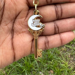 May include: Gold-colored key chain with a key-shaped pendant. The pendant features a white crescent moon with the words "UNLOCK YOUR POTENTIAL" on a banner. The key chain is held against a background of green grass.