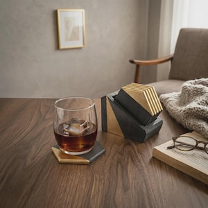Op de afbeelding: Een glas amberkleurige vloeistof met ijs staat op een zeshoekige onderzetter. Een set geometrische onderzetters in een houder, met zwarte en gouden accenten, staat naast het glas. Een boek en een bril liggen op tafel.