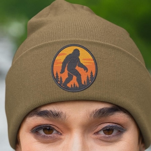 May include: Olive green beanie featuring a circular patch with a Bigfoot silhouette against a sunset backdrop. The patch includes trees and a yellow and orange sky. The beanie is made of a soft material.