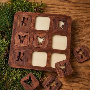 May include: A wooden tic-tac-toe game with a dark brown frame and light wood squares. The game pieces are small, square, and feature cut-out designs of butterflies and figures. The game is set on a wooden surface with moss and pine needles.