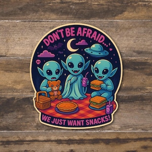 May include: A sticker featuring three blue aliens having a picnic. The aliens are gathered around a red and white checkered blanket, with a teddy bear, pie, sandwiches, and drinks. The text reads "DON'T BE AFRAID... WE JUST WANT SNACKS!"