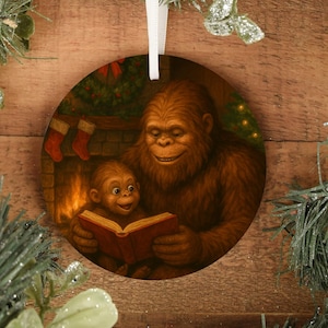 Könnte beinhalten: Runder Weihnachtsschmuck mit einem Sasquatch, der einem kleineren Sasquatch ein Buch vorliest. Die Illustration zeigt einen Kamin mit Strümpfen und einem Weihnachtsbaum. Der Schmuck hat ein weißes Band zum Aufhängen.
