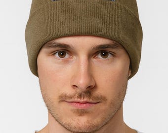 May include: Olive green beanie with a black and red embroidered graphic. The beanie is made of a soft, knitted material and has a folded cuff. The graphic is a stylized creature with red eyes.