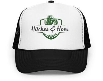 May include: A black and white trucker hat with a white front panel. The front features a green tractor graphic and the words "Hitches & Hoes" in a stylized font. The hat has a curved brim and a mesh back.