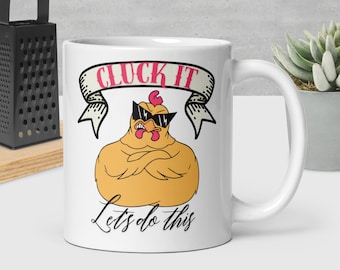 May include: White ceramic mug featuring a cartoon chicken wearing sunglasses with the text "CLUCK IT" in a banner above the chicken and "Lets do this" below. The mug has a curved handle and is on a light surface.
