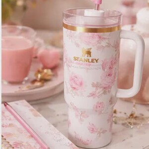May include: A white Stanley tumbler with a floral rose pattern and a pink bow straw topper. The tumbler has a gold rim and a white handle. A pink pen and notebook are in the foreground.