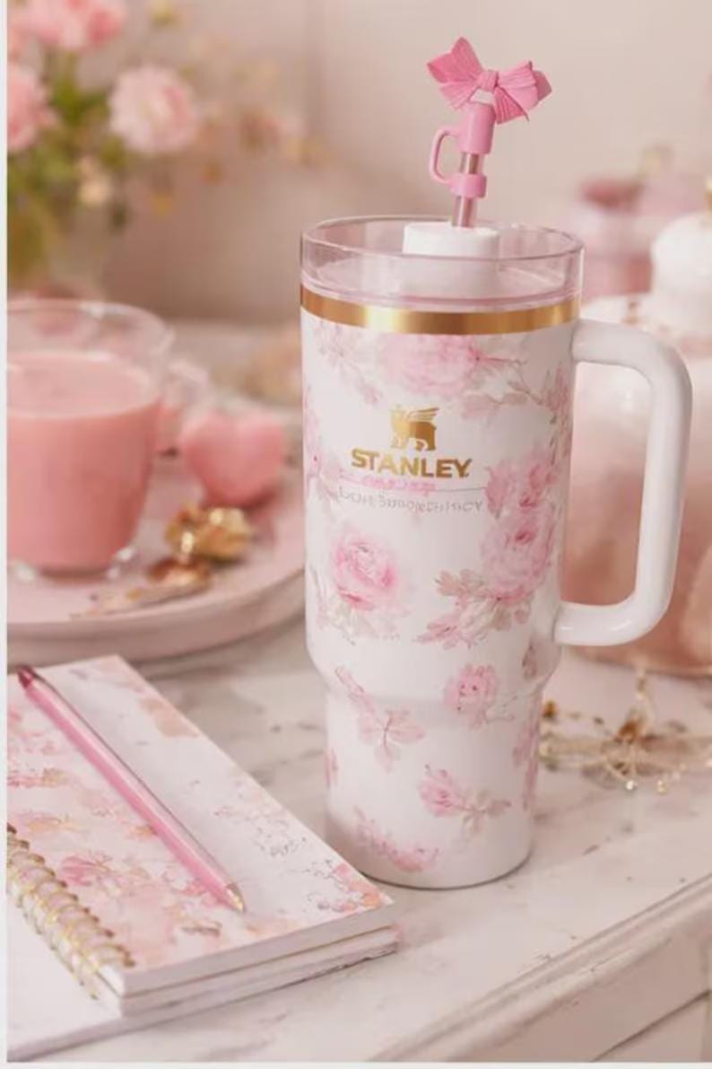 May include: A white Stanley tumbler with a floral rose pattern and a pink bow straw topper. The tumbler has a gold rim and a white handle. A pink pen and notebook are in the foreground.
