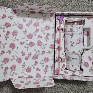 May include: A white Stanley Quencher H2.0 tumbler with a pink floral pattern, presented in a matching floral-printed box. The tumbler has a handle, a straw, and a lid. The box and tumbler feature pink roses.