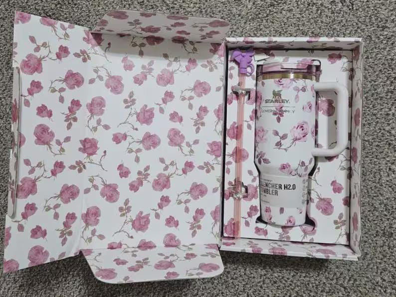 May include: A white Stanley Quencher H2.0 tumbler with a pink floral pattern, presented in a matching floral-printed box. The tumbler has a handle, a straw, and a lid. The box and tumbler feature pink roses.