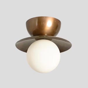 May include: A modern ceiling light fixture with a bronze-colored bowl-shaped top, a dark gray disc, and a white spherical light. The fixture has a minimalist design and is suitable for various interior styles.