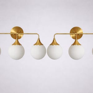 May include: Two matching light fixtures with a gold finish. Each has a round gold backplate, a horizontal gold bar, and three white globe lights with gold accents. Designed for wall mounting, these lights are ideal for a bathroom.