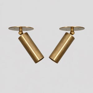 May include: Two brass-coloured, cylindrical spotlights with a circular base and a ball joint. The spotlights are angled and appear to be made of metal. The background is a solid, neutral colour.