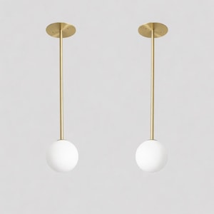 May include: Two pendant lights featuring a gold finish and white globe shades. Each light hangs from a circular ceiling mount. The design is minimalist and modern, suitable for various interior styles.