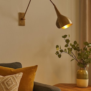 Op de afbeelding: Een messing wandlamp met een verstelbare arm en een conische kap. De lamp bevindt zich boven een donkergrijze bank met gele en patroon kussens. Een houten bijzettafel bevat een vaas met groen.