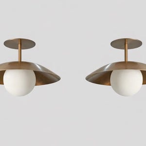 Könnte beinhalten: Zwei moderne Deckenleuchten mit Messing-Finish. Jede Leuchte hat einen kreisförmigen Sockel, einen zentralen Stiel, einen breiten, nach unten gerichteten Schirm und eine kugelförmige weiße Glaskugel. Vor einem schlichten weißen Hintergrund.