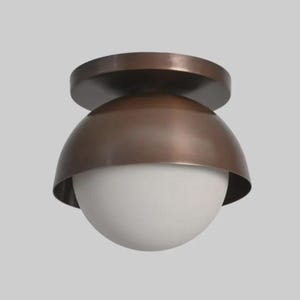 May include: A bronze-colored flush mount ceiling light fixture. The fixture has a round, flat base, a curved, dome-shaped shade, and a white, spherical glass globe. The light is designed for ceiling installation and provides ambient lighting.
