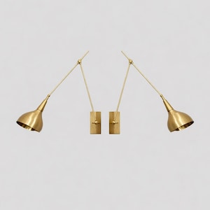 May include: Two brass-toned wall sconces with adjustable arms and bell-shaped shades. The sconces are mounted on rectangular bases and have a minimalist, modern design. The light fixtures are set against a plain white background.