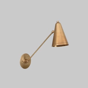 Peut inclure: Une applique murale en laiton avec un abat-jour conique et un bras réglable. La lampe a une base circulaire et une série de petits trous autour du bas de l'abat-jour. La finition est dorée et métallique.