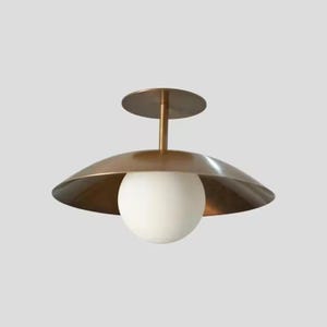 May include: A modern ceiling light fixture with a brass-colored shade and a white globe light. The fixture has a circular base and a stem connecting the shade to the base. The shade is a wide, shallow bowl shape.