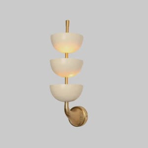 May include: A modern wall sconce with three off-white, bowl-shaped shades stacked vertically. The shades are connected by brass rods and mounted on a curved brass arm. The sconce emits a warm, inviting light.