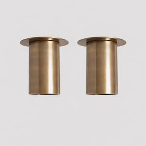 May include: Two cylindrical brass objects with a flat, circular top. The objects have a warm, metallic finish and are set against a plain white background. The design is minimalist and modern.