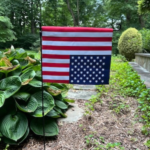 May include: A small American flag garden flag with red and white stripes and a blue square with white stars. The flag is mounted on a black metal stand and displayed in a garden setting with green foliage and mulch.