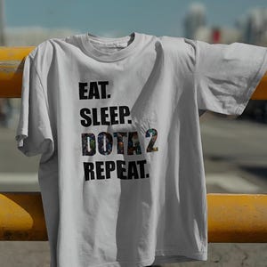 Puede incluir: Una camiseta blanca con el texto "EAT. SLEEP. DOTA 2. REPEAT." en letras negras. El texto "DOTA 2" está relleno con un gráfico colorido. La camiseta está sobre una barra de metal amarilla, con un cielo azul de fondo.