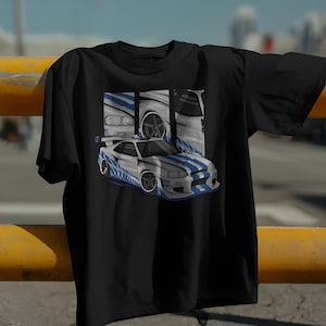 May include: Black t-shirt featuring a graphic of a white and blue sports car. The design includes a detailed illustration of the car's exterior and wheels, with blue stripes. The shirt is hanging on a yellow metal bar.
