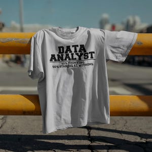 Pode incluir: Uma camiseta cinza clara com as palavras "DATA ANALYST" em letras pretas em negrito. Abaixo, lê-se "50% THINKING 50% STARING AT NOTHING". A camiseta está sobre uma barreira metálica amarela.