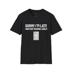 Puede incluir: Camiseta negra con el texto blanco "SORRY I'M LATE I WAS BUSY READING LABELS". Un pequeño gráfico de una etiqueta de información nutricional está debajo del texto. La camiseta es de cuello redondo.