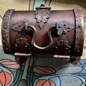 May include: A small, barrel-shaped, copper-colored chest with ornate leaf-shaped accents and a curved handle. The chest has a hinged lid with a clasp and decorative metalwork. The chest is supported by small feet.