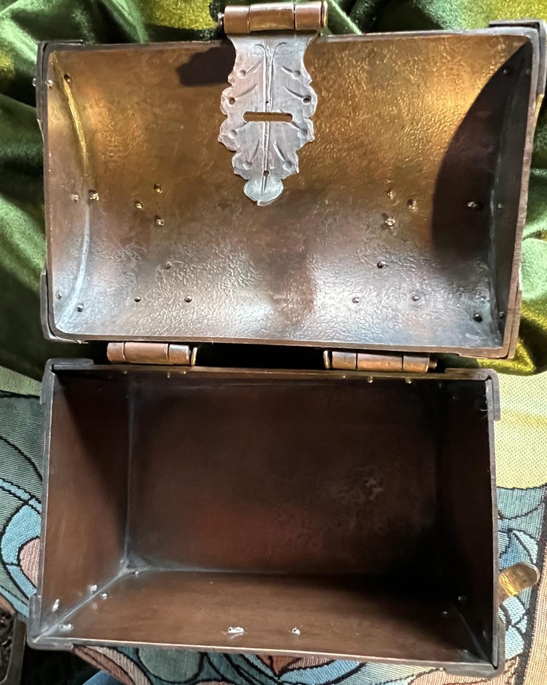 May include: An open, antique-style metal box with a hinged lid. The box is a dark bronze color with a decorative silver-colored clasp. The interior of the box is empty. The box is likely used for storage or display.