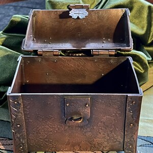 May include: An open, antique-style metal chest with a hinged lid. The chest is a dark, mottled brown with decorative floral accents on the sides and feet. The lid features a decorative clasp. The interior is empty.