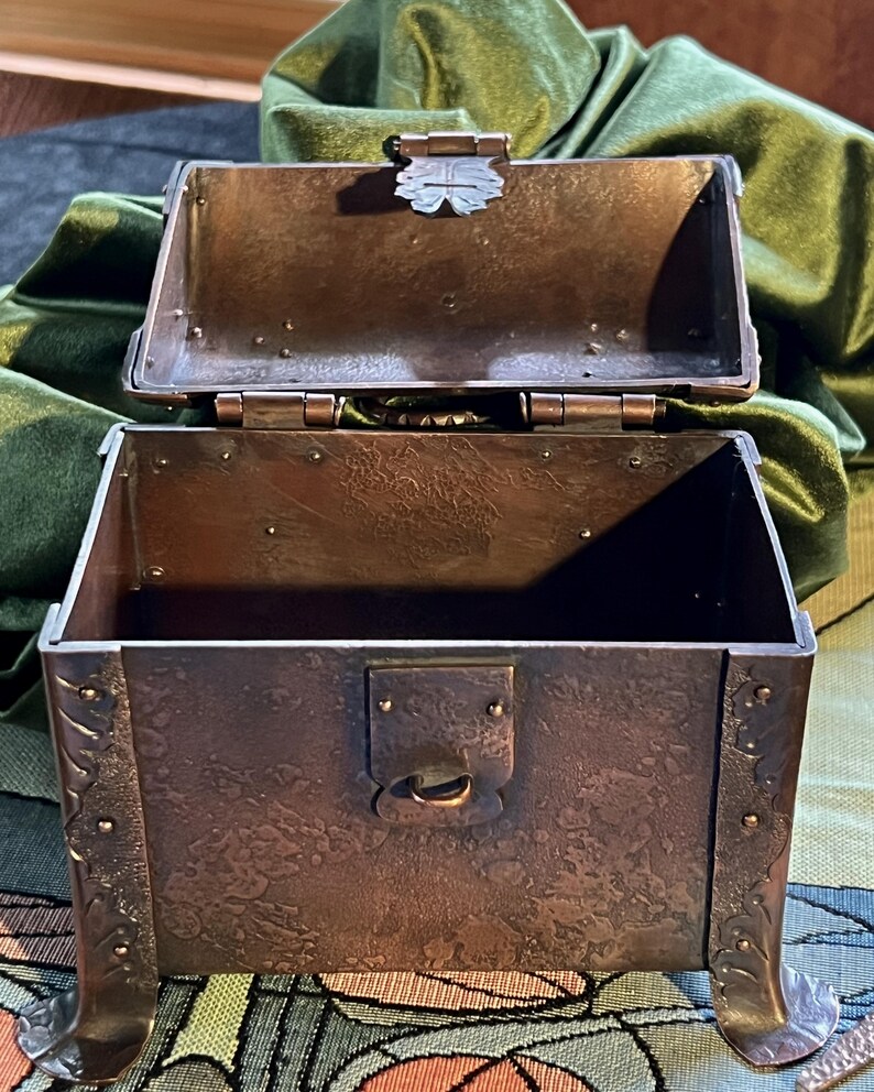 May include: An open, antique-style metal chest with a hinged lid. The chest is a dark, mottled brown with decorative floral accents on the sides and feet. The lid features a decorative clasp. The interior is empty.