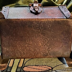 May include: A handcrafted copper box with a textured surface and decorative accents. The box features a hinged lid with a clasp and is supported by four curved feet. The overall design is rustic and vintage.