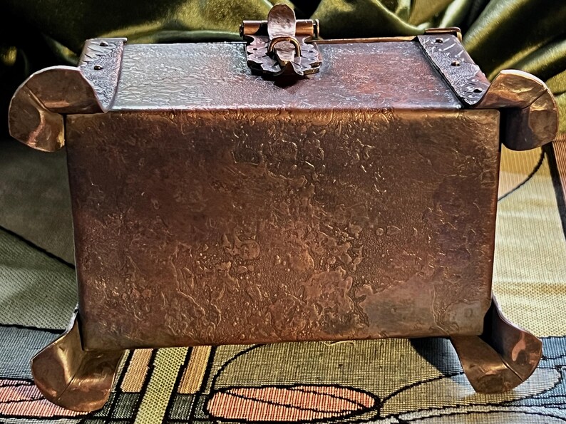 May include: A handcrafted copper box with a textured surface and decorative accents. The box features a hinged lid with a clasp and is supported by four curved feet. The overall design is rustic and vintage.