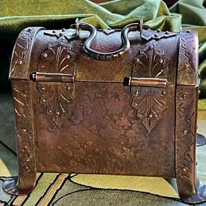 May include: A small, antique copper chest with ornate leaf and stud detailing. The chest has a curved top, a dark metal handle, and decorative metal accents. The chest is raised on four small feet and has a hinged lid.