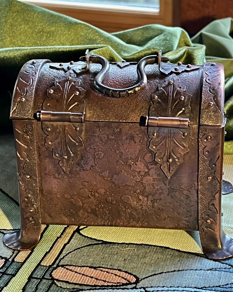 May include: A small, antique copper chest with ornate leaf and stud detailing. The chest has a curved top, a dark metal handle, and decorative metal accents. The chest is raised on four small feet and has a hinged lid.