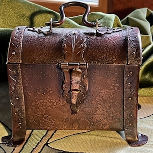 May include: A small, antique-style chest with a copper finish. The chest features decorative metalwork, including a handle, hinges, and a clasp. The chest has four small feet and is designed to resemble a miniature treasure chest.