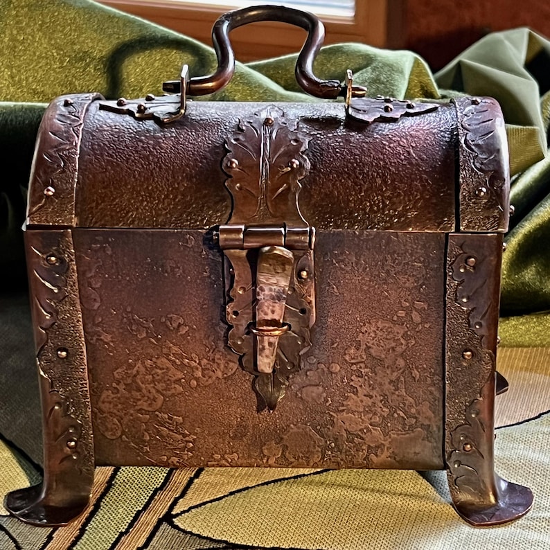 May include: A small, antique-style chest with a copper finish. The chest features decorative metalwork, including a handle, hinges, and a clasp. The chest has four small feet and is designed to resemble a miniature treasure chest.