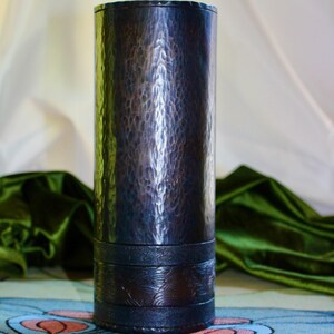 May include: A tall, dark metal vase with a hammered texture. The vase has a cylindrical shape with a decorative band near the base. The color is a dark, metallic bronze with a slightly reflective surface.